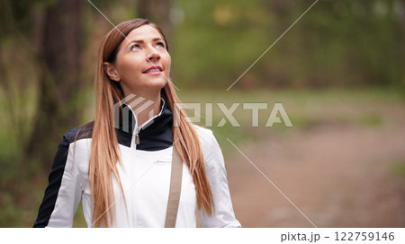 Young woman with long hair, wearing white jacket, looking up the trees while walking in forest. Space for text right side 122759146