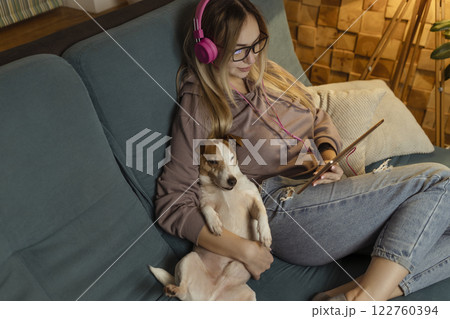 A girl on the couch lies with a dog in her laptop and listens to music through headphones. Freelancing and communication via the Internet A girl on the couch lies with a dog in her laptop and listens to music through headphones. Freelancing and communication via the Internet 122760394