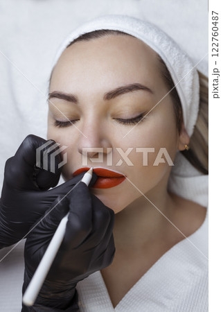 Young woman with permanent makeup on her lips in a cosmetologists salon. Permanent makeup tattoo. draw a line with a White lip pencil 122760487