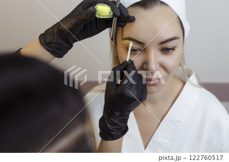 The hand of a make-up artist in black gloves applies a yellow eyebrow paste to the eyebrow. Professional stylish permanent makeup, eyebrow tinting. Close up. The hand of a make-up artist in black gloves applies a yellow eyebrow paste to the eyebrow. Professional stylish permanent makeup, eyebrow tinting. Close up. 122760517