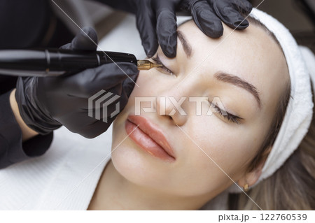 Close-up of a woman, in a beauty salon with her eyes closed, a cosmetologist makes permanent eyeliner makeup. Professional in black gloves and with a special too Close-up of a woman, in a beauty salon with her eyes closed, a cosmetologist makes permanent eyeliner makeup. Professional in black gloves and with a special too 122760539