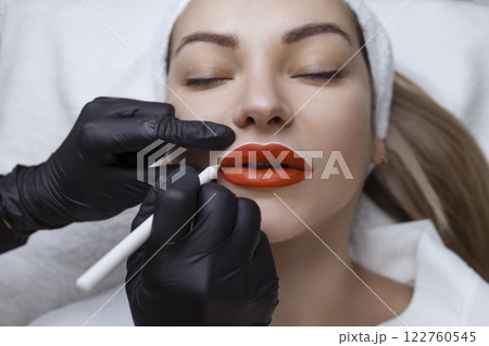 Young woman with permanent makeup on her lips in a cosmetologists salon. Permanent makeup tattoo. draw a line with a White lip pencil Young woman with permanent makeup on her lips in a cosmetologists salon. Permanent makeup tattoo. draw a line with a White lip pencil 122760545