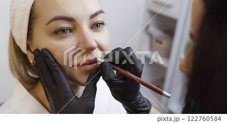 Young woman with permanent makeup on her lips in a cosmetologists salon. Permanent makeup tattoo. draw a line with a red lip pencil 122760584