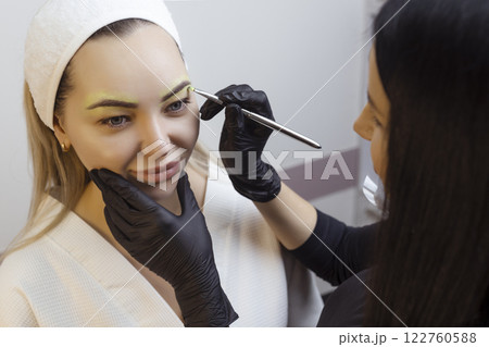 The hand of a make-up artist in black gloves applies a yellow eyebrow paste to the eyebrow. Professional stylish permanent makeup, eyebrow tinting. Close up. 122760588