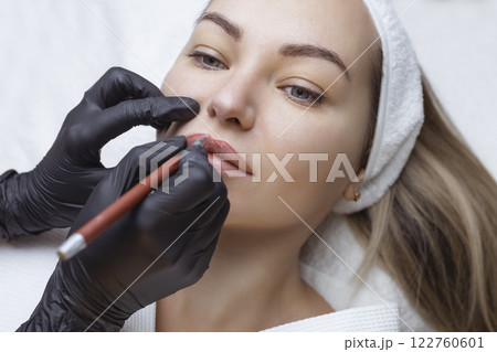 Young woman with permanent makeup on her lips in a cosmetologists salon. Permanent makeup tattoo. draw a line with a red lip pencil Young woman with permanent makeup on her lips in a cosmetologists salon. Permanent makeup tattoo. draw a line with a red lip pencil 122760601