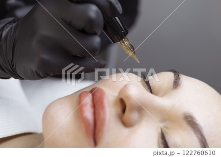 Close-up of a woman, in a beauty salon with her eyes closed, a cosmetologist makes permanent eyeliner makeup. Professional in black gloves and with a special too Close-up of a woman, in a beauty salon with her eyes closed, a cosmetologist makes permanent eyeliner makeup. Professional in black gloves and with a special too 122760610