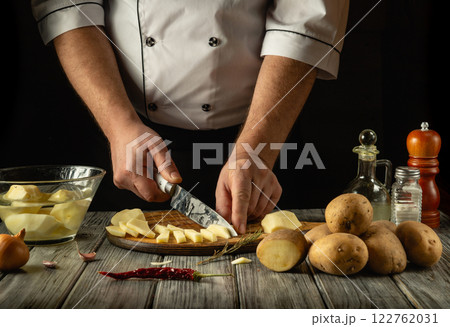 A skilled chef engages in culinary preparation, chopping fresh potatoes with precision. Various ingredients are arranged artistically on a wooden countertop, blending rustic charm and gastronomy A skilled chef engages in culinary preparation, chopping fresh potatoes with precision. Various ingredients are arranged artistically on a wooden countertop, blending rustic charm and gastronomy 122762031