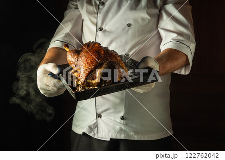 In a dimly lit kitchen, a skilled chef proudly holds a roasted duck on a tray. The rich golden-brown skin glistens, inviting the senses to indulge in this exquisite culinary creation In a dimly lit kitchen, a skilled chef proudly holds a roasted duck on a tray. The rich golden-brown skin glistens, inviting the senses to indulge in this exquisite culinary creation 122762042