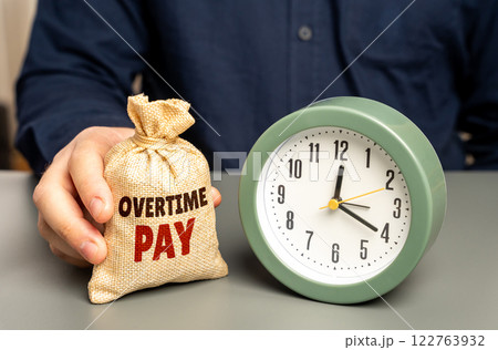 A man holds a money bag with overtime pay near a clock. Concept of additional compensation payment and incentive to work overtime. Wage increases, bonuses, extra hours, payroll benefits for hard work 122763932