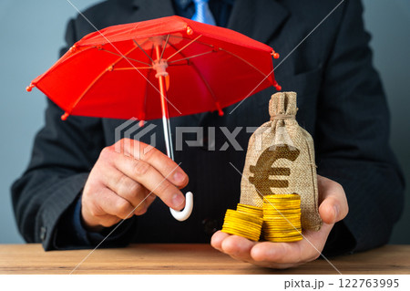 Businessman holding euro money bag with coins under red umbrella. Capital preservation. Savings and deposits insurance concept. Businessman holding euro money bag with coins under red umbrella. Capital preservation. Savings and deposits insurance concept. 122763995