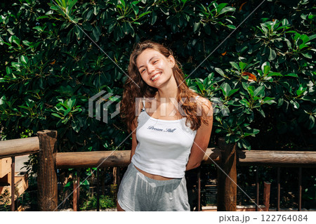 Smiling young woman leans on a wooden railing surrounded by lush greenery, enjoying a sunny day 122764084