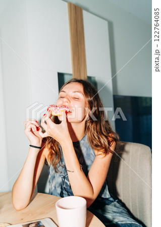 A cheerful young woman bites into a pink-frosted donut topped with sprinkles, enjoying a sweet treat 122764085