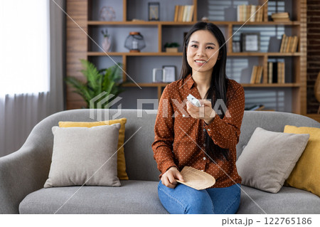 Joyful woman holding air conditioner remote control, Asian woman sitting at home on sofa in living room, switching cooling programs on air conditioner, smart home system. 122765186