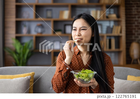 Asian woman eating salad with dissatisfied expression, contemplating diet. Seated on couch in contemporary interior setting, representing healthy lifestyle choices. Concept of wellness journey. 122765191