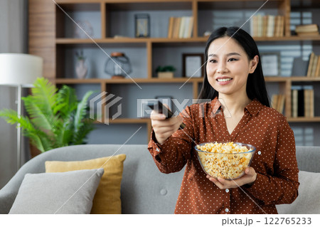 Asian woman holding bowl of popcorn and TV remote, enjoying movie night at home. Smiling, relaxed atmosphere, showcasing leisure and entertainment in modern living room setting. 122765233