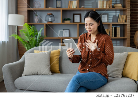 Asian woman holding tablet sits on gray couch in cozy living room. She appears thoughtful, possibly engaged in video call or online communication, wearing casual brown polka dot shirt. Asian woman holding tablet sits on gray couch in cozy living room. She appears thoughtful, possibly engaged in video call or online communication, wearing casual brown polka dot shirt. 122765237