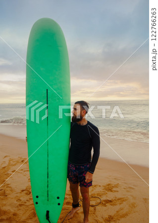 Young  bearded man with surfboard standing near a beach. Man with surfing board outdoors on a summer day. 122766263