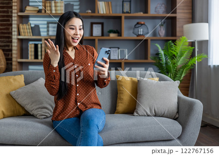 Asian woman celebrates online success, excited expression, holding smartphone. Sitting on cozy couch, modern living room environment. Positive vibes, home comfort, digital connection theme. 122767156