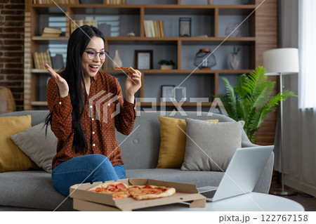 Asian woman with glasses sits on couch holding pizza slice while video chatting on laptop. She looks happy and surprised, enjoying leisure time in comfortable home environment. Asian woman with glasses sits on couch holding pizza slice while video chatting on laptop. She looks happy and surprised, enjoying leisure time in comfortable home environment. 122767158