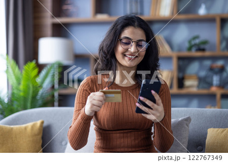 Close-up photo of a young smiling Muslim girl at home, sitting on the sofa, holding a credit card and using a mobile phone. 122767349