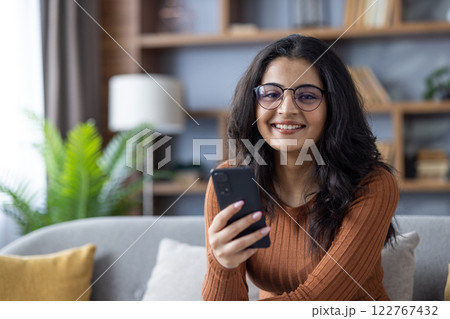 Close-up portrait of young Muslim woman sitting on sofa at home, holding phone in hand and looking at camera. Close-up portrait of young Muslim woman sitting on sofa at home, holding phone in hand and looking at camera. 122767432