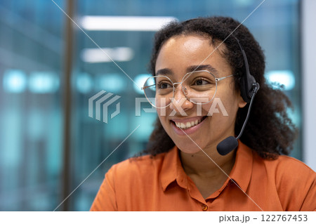 Young successful support worker at workplace inside office with headset, woman smiling close up looking to the side. Young successful support worker at workplace inside office with headset, woman smiling close up looking to the side. 122767453