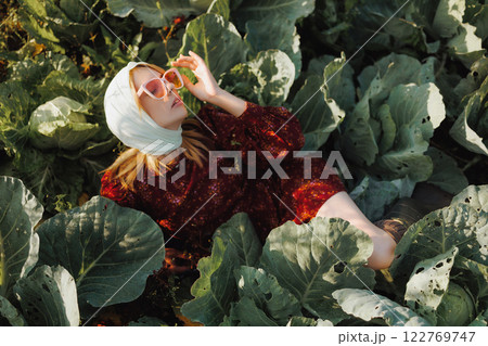 A woman model in a red dress and sunglasses is posing lying on a cabbage field. A woman model in a red dress and sunglasses is posing lying on a cabbage field. 122769747
