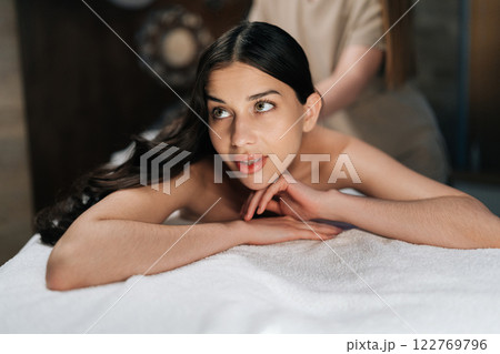 Close-up face of attractive female client getting relaxing back massage from professional massage therapist while lying resting on table, experiencing therapeutic wellness at luxurious spa setting. 122769796