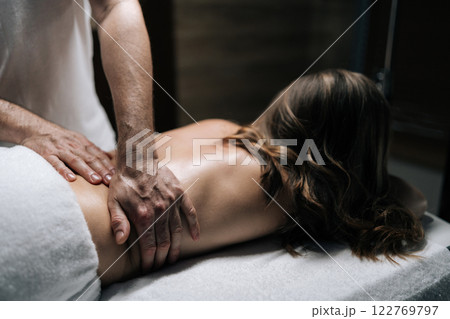 Close-up hands of masseur male performing deep tissue technique on female lower back, targeting muscle tension and promoting wellness during bodywork session in beauty parlor with dark interior. Close-up hands of masseur male performing deep tissue technique on female lower back, targeting muscle tension and promoting wellness during bodywork session in beauty parlor with dark interior. 122769797