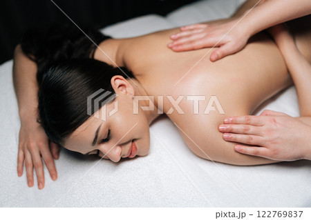 High-angle view of serene young woman lying on massage table with closed eyes during professional relaxing massage at spa salon. Skilled massage therapist performing soothing back treatment. High-angle view of serene young woman lying on massage table with closed eyes during professional relaxing massage at spa salon. Skilled massage therapist performing soothing back treatment. 122769837