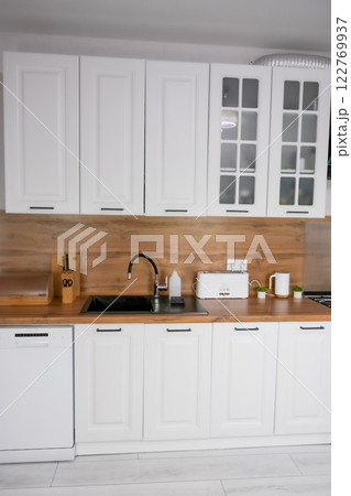 Horizontal photo of a modern kitchen in white and wooden style, in the center is a black kitchen sink, on a wooden countertop there are white wooden appliances for storing and preparing food. Modern 122769937