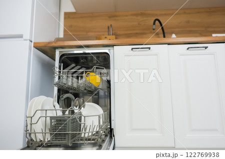 An open dishwasher with a dish drawer pulled out close-up on the left, it contains clean dishes and cutlery against the background of a white wooden kitchen. Comfort concept, dishwashing. High quality An open dishwasher with a dish drawer pulled out close-up on the left, it contains clean dishes and cutlery against the background of a white wooden kitchen. Comfort concept, dishwashing. High quality 122769938
