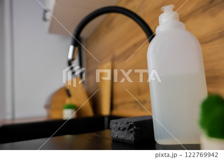 Close-up of a white bottle of dishwashing soap, a black dishwashing sponge lying next to it and a potted plant against a background of a black granite sink and a flexible high faucet and a white Close-up of a white bottle of dishwashing soap, a black dishwashing sponge lying next to it and a potted plant against a background of a black granite sink and a flexible high faucet and a white 122769942