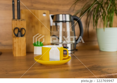 A yellow cup of tea stands on a yellow saucer and on the saucer lies a tea bag on a wooden table against the background of a teapot, a plant and kitchen utensils. The concept of tea drinking, break 122769947
