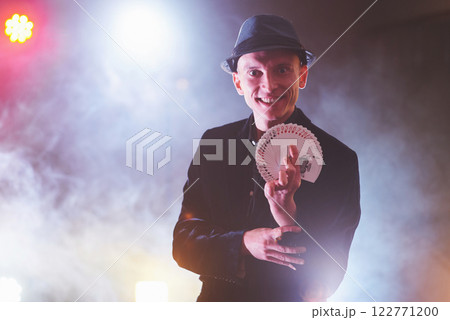 Magician showing trick with playing cards. Magic or dexterity, circus, gambling. Prestidigitator in dark room with fog Magician showing trick with playing cards. Magic or dexterity, circus, gambling. Prestidigitator in dark room with fog 122771200