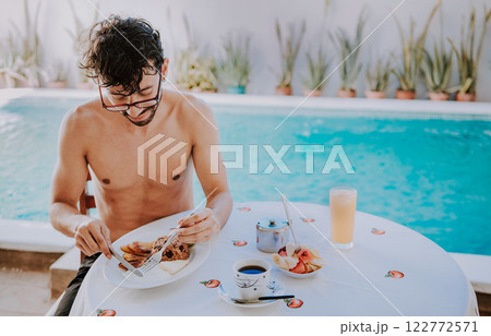 Young man on vacation in hotel having breakfast near swimming pool. Breakfast near the swimming pool. Person having breakfast in the hotel with pool in the background Young man on vacation in hotel having breakfast near swimming pool. Breakfast near the swimming pool. Person having breakfast in the hotel with pool in the background 122772571