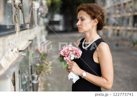Sad woman in black dress with bouquet of flowers and handkerchief in hands, stands in cemetery 122773741