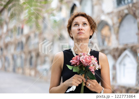 Upset woman in black dress, who lost loved one, brings flowers to grave 122773864