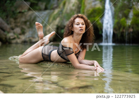 Woman in mesh black bikini sits on rock on shore of tropical waterfall Woman in mesh black bikini sits on rock on shore of tropical waterfall 122773925