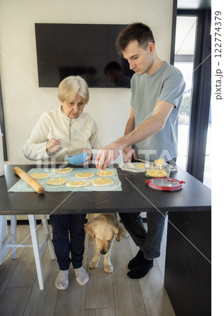 Elderly woman and young man preparing homemade pastries in a modern kitchen, spreading cheese on dough. A Golden Retriever stands under the table, waiting for food. 122774379