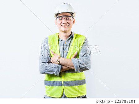 Portrait of handsome and smiling engineer with crossed arms isolated. Portrait of a young cheerful engineer in helmet and vest isolated. 122785101