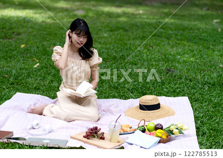 Young woman is sitting on a cloth on the grass with food and snacks Young woman is sitting on a cloth on the grass with food and snacks 122785513