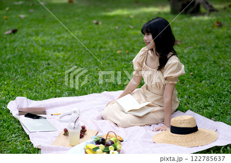 Young woman is sitting on a cloth on the grass with food and snacks 122785637