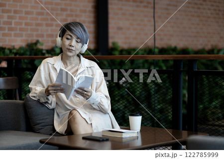 Elder woman sitting on sofa wearing headphones reading a book Elder woman sitting on sofa wearing headphones reading a book 122785999