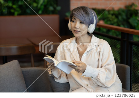 Elder woman sitting on sofa  wearing headphones reading a book 122786025
