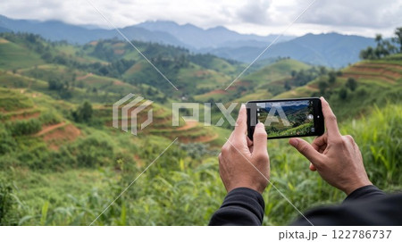 Hands holding smartphone capturing stunning mountain landscape with terraced rice fields. Travel photography of scenic Asian agricultural vista with rolling hills and cloudy sky Hands holding smartphone capturing stunning mountain landscape with terraced rice fields. Travel photography of scenic Asian agricultural vista with rolling hills and cloudy sky 122786737