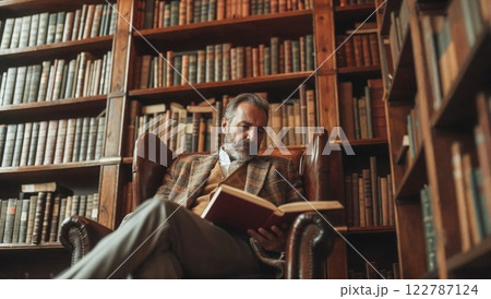 distinguished man in a tweed jacket sits in a classic leather armchair, engrossed in a book, surrounded by towering wooden bookshelves filled with antique books in a warm-lit library 122787124
