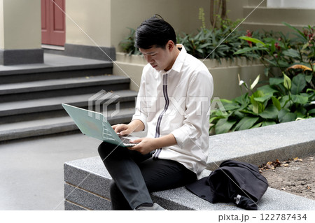 Businessman in casual clothes using laptop outdoors in walkway and garden seating area Businessman in casual clothes using laptop outdoors in walkway and garden seating area 122787434