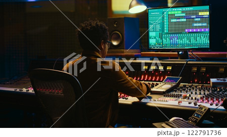 African american audio engineer uses compressor and mixer in professional studio, changing volume level and sound settings on a recording. Music production in control room. Camera A. 122791736