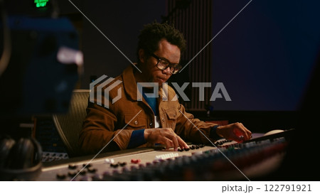 Music producer using audio mixer in professional studio, adjusting volume levels while pressing buttons and sliders on control desk. Sound designer works with digital audio software. Camera A. 122791921